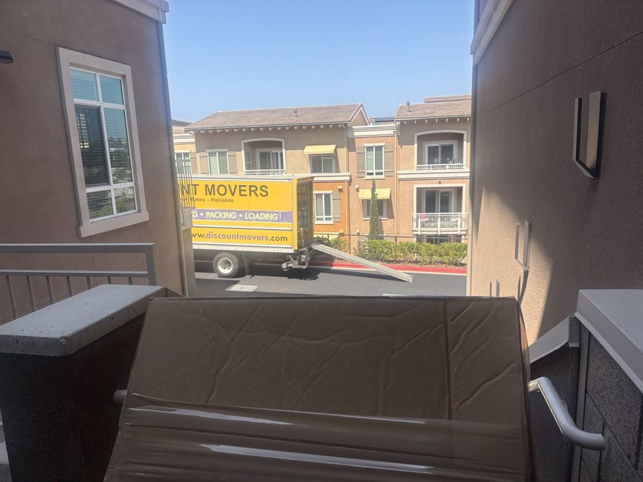 A moving box carried through an apartment breezeway toward the Discount Movers truck parked outside in Carmel Valley