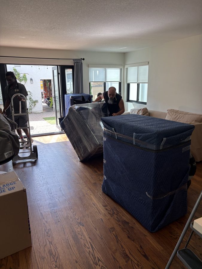 Blanket-wrapped sofa and shrink-wrapped chest of drawers ready to carry out of a Coronado living room, two Discount Movers crew members behind them
