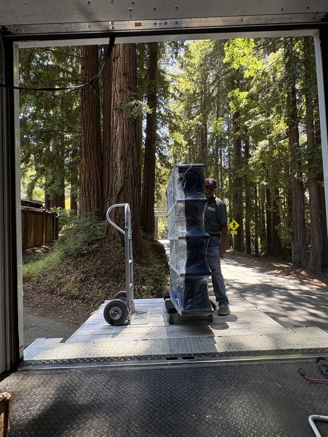 Blanket-wrapped and shrink-wrapped furniture on a dolly at the truck liftgate, redwoods behind, at a Northern California delivery