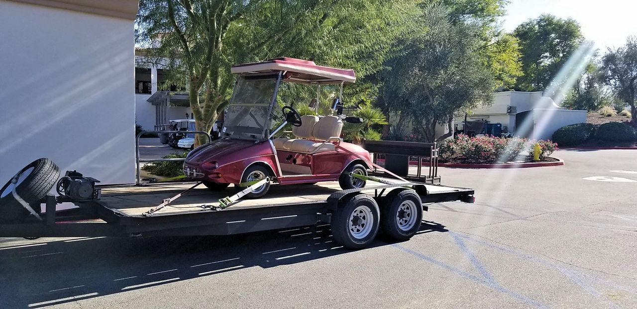 Custom red golf cart ratchet-strapped at the frame to a Discount Movers flatbed trailer outside a country club clubhouse