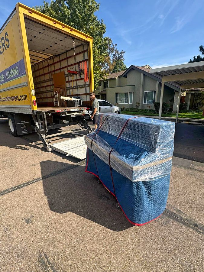 Blanket-wrapped and shrink-wrapped piano strapped to a dolly at the open liftgate of a Discount Movers truck