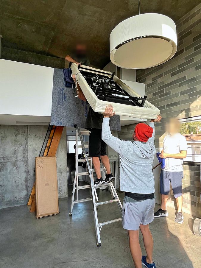 Discount Movers crew hoisting an adjustable bed base up to a loft level from a ladder, with a second mover guiding from above