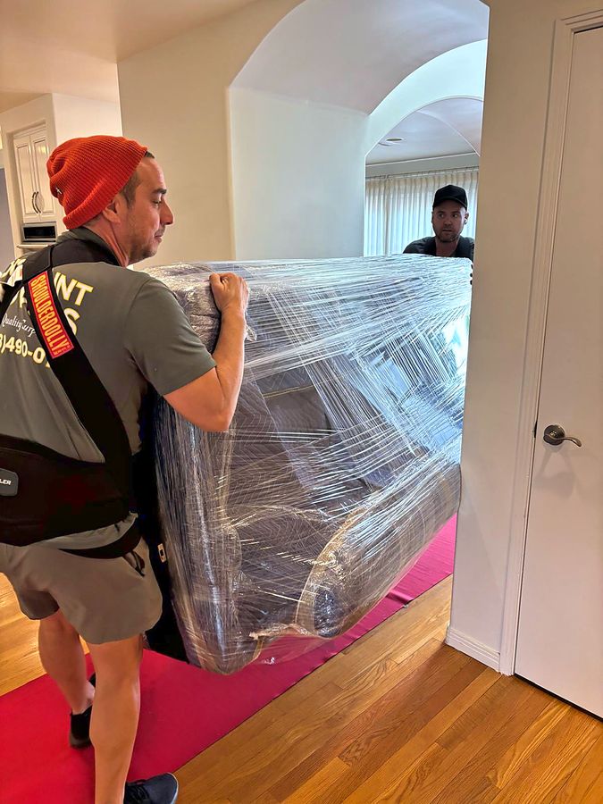 Two Discount Movers crew members carrying a shrink-wrapped sofa across a protective floor runner inside a home