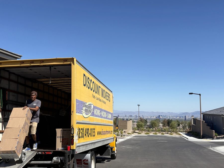 Discount Movers box truck with its ramp down at a Las Vegas delivery, a crew member wheeling boxes off the liftgate, the Las Vegas Strip on the horizon