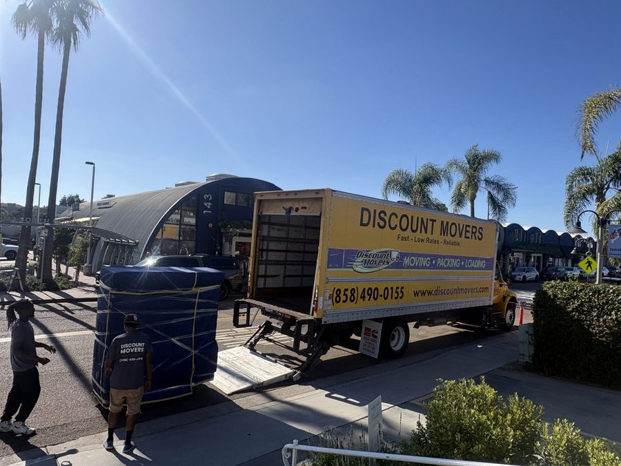 Discount Movers truck with the liftgate down on Cedros Avenue in Solana Beach, two crew members guiding a blanket-wrapped commercial refrigerated display case on a dolly