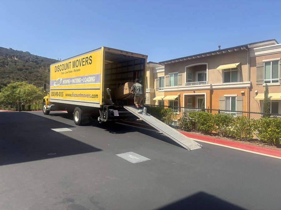 Discount Movers truck with the ramp down at a Carmel Valley apartment complex, a crew member carrying a wrapped piece up into the truck